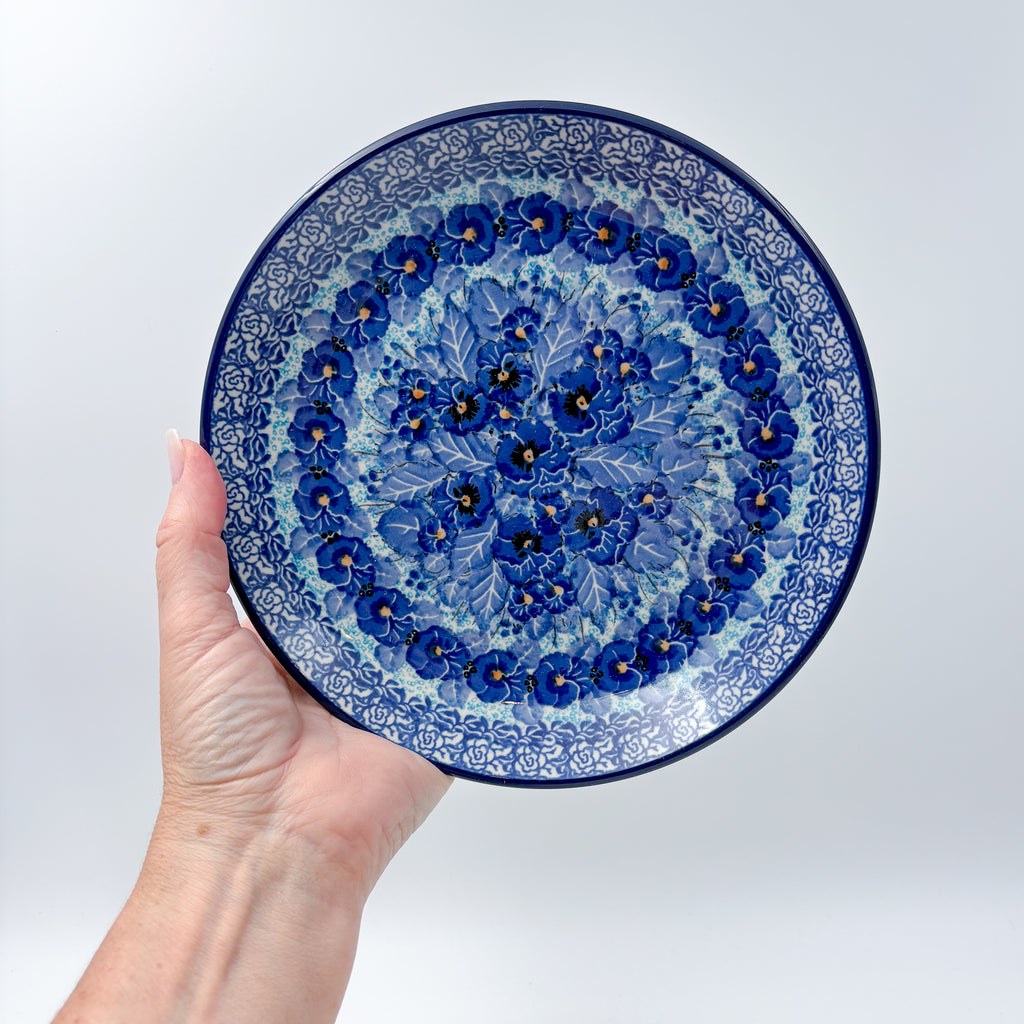 UNIKAT Deep Winter plate held in hand, front view showing blue pansy cluster and rose border