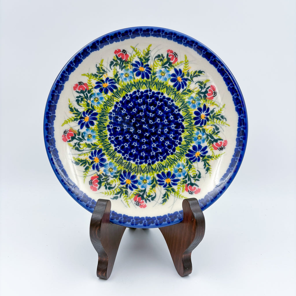 UNIKAT Meadow Breeze plate, front view displayed on wooden stand showing blue wildflower ring around navy center