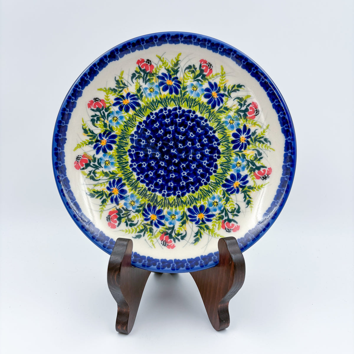 UNIKAT Meadow Breeze plate, front view displayed on wooden stand showing blue wildflower ring around navy center
