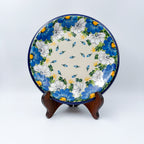 UNIKAT Blue Petal Dance plate, front view displayed on wooden stand showing blue and white daisy ring