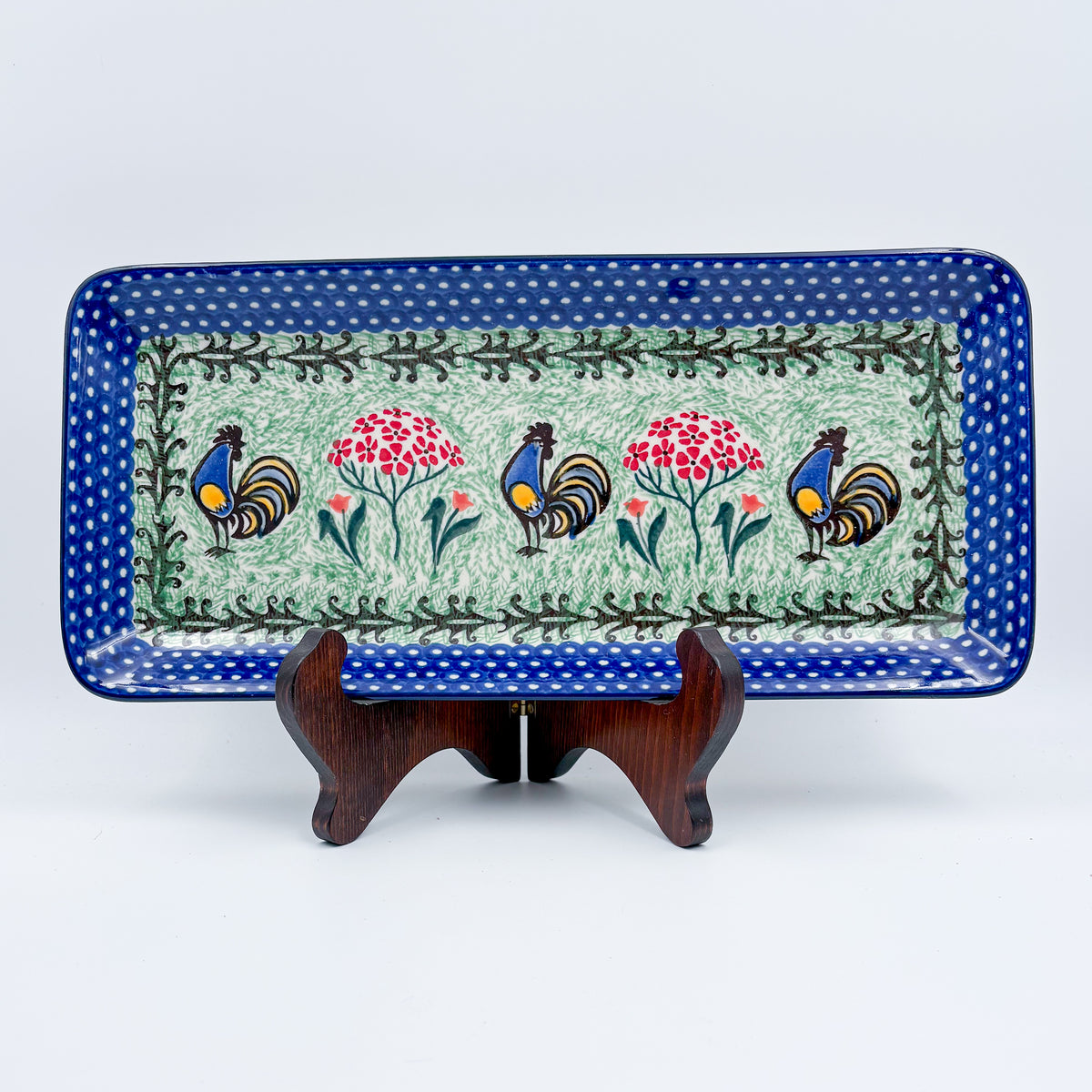 UNIKAT Rooster Dance rectangular platter, front view displayed on wooden stand showing roosters and pom-pom flowers