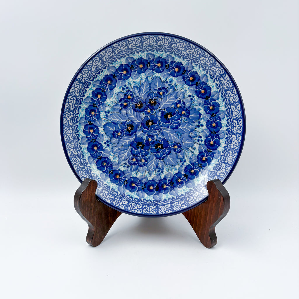 UNIKAT Deep Winter plate, front view displayed on wooden stand showing blue pansy cluster and rose border
