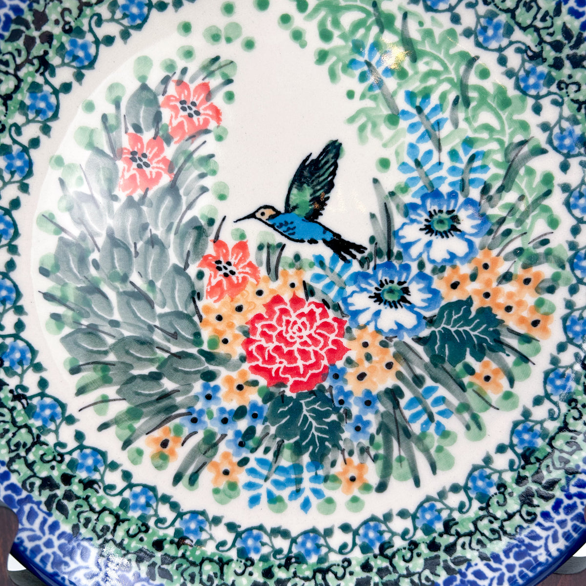 UNIKAT Hummingbird Meadow plate, close-up top-down view of hummingbird among red and blue flowers