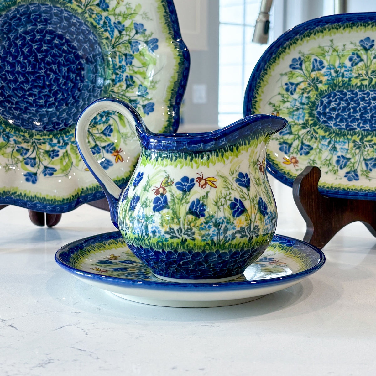 A Polish pottery gravy boat and saucer painted with blue flowers, with matching platters on stands behind it