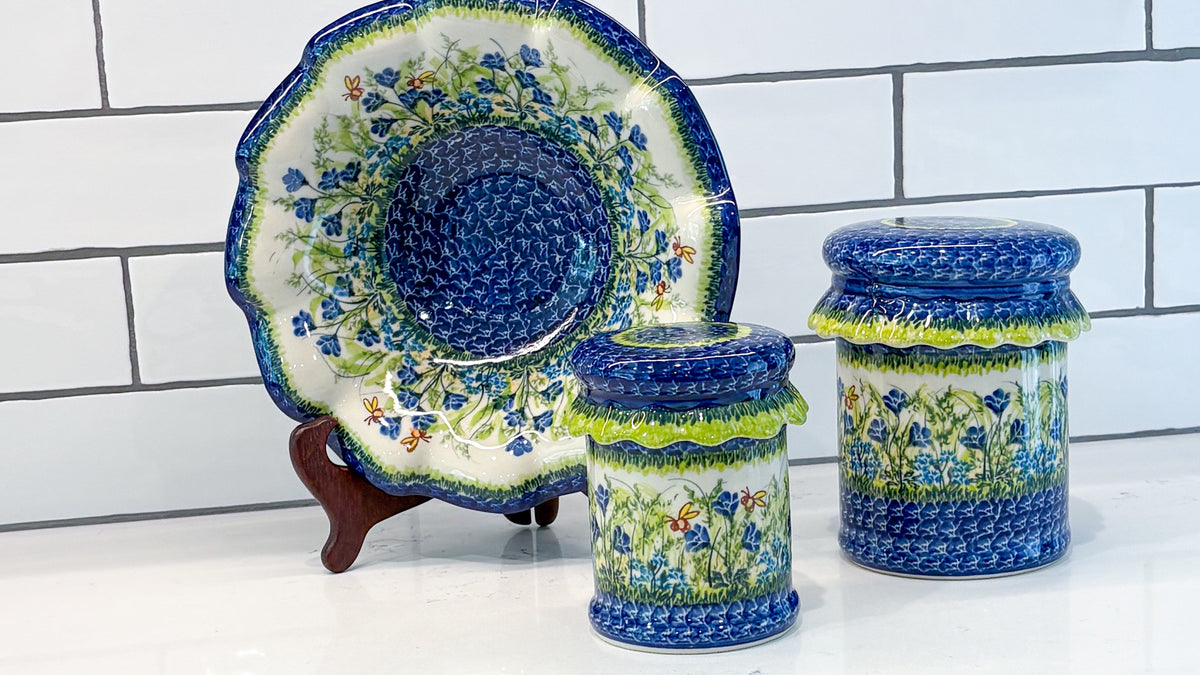 A ruffled Polish pottery platter on a stand beside two lidded jars, painted with blue flowers, against white kitchen tile
