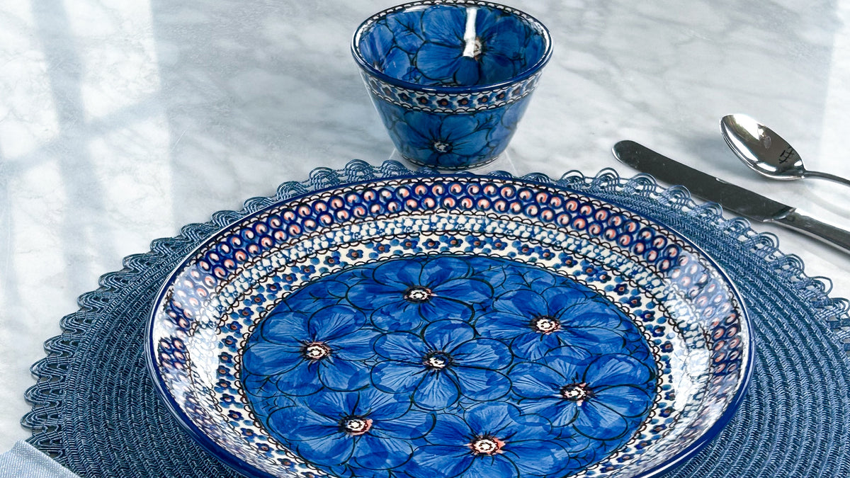 A Polish pottery dinner plate and small bowl painted with large blue flowers, set on a blue placemat on a marble table