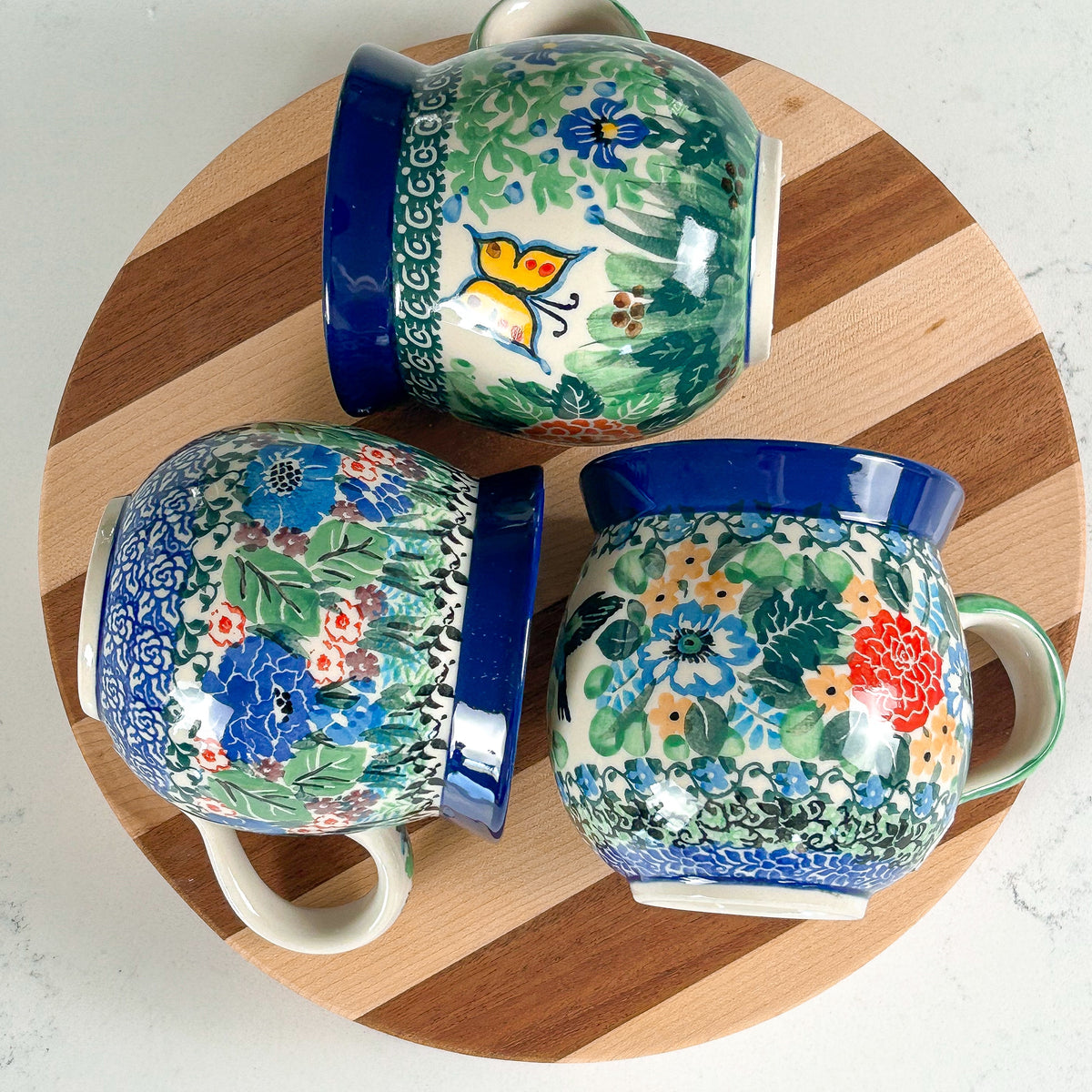 Three Polish pottery mugs in different floral patterns, one with a yellow butterfly, on a round wooden board